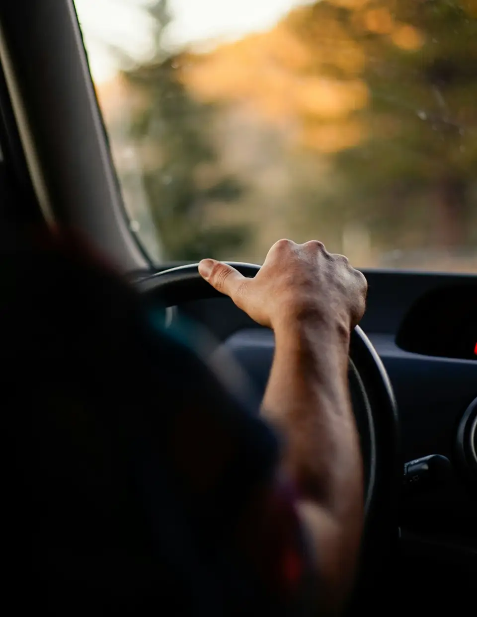 A hand resting on a car steering wheel, dashboard softly lit by warm late-afternoon light through the windshield.