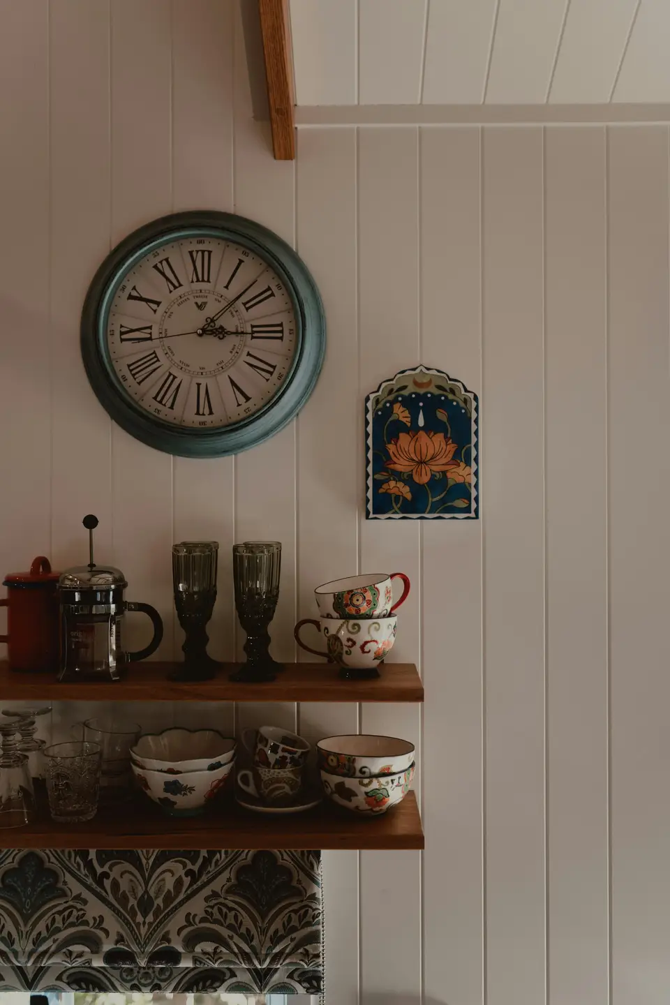 A vintage wall clock above open shelves holding mismatched mugs and a French press, in a warmly lit kitchen.