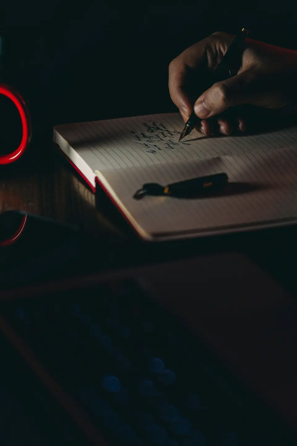 A hand writing in a notebook by lamplight in the evening, pen mid-word.