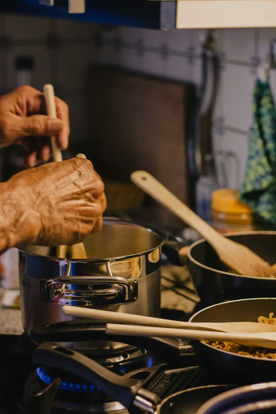 A hand stirring a pot on a home stovetop, with a second pan and a wooden spoon beside it in warm evening light.