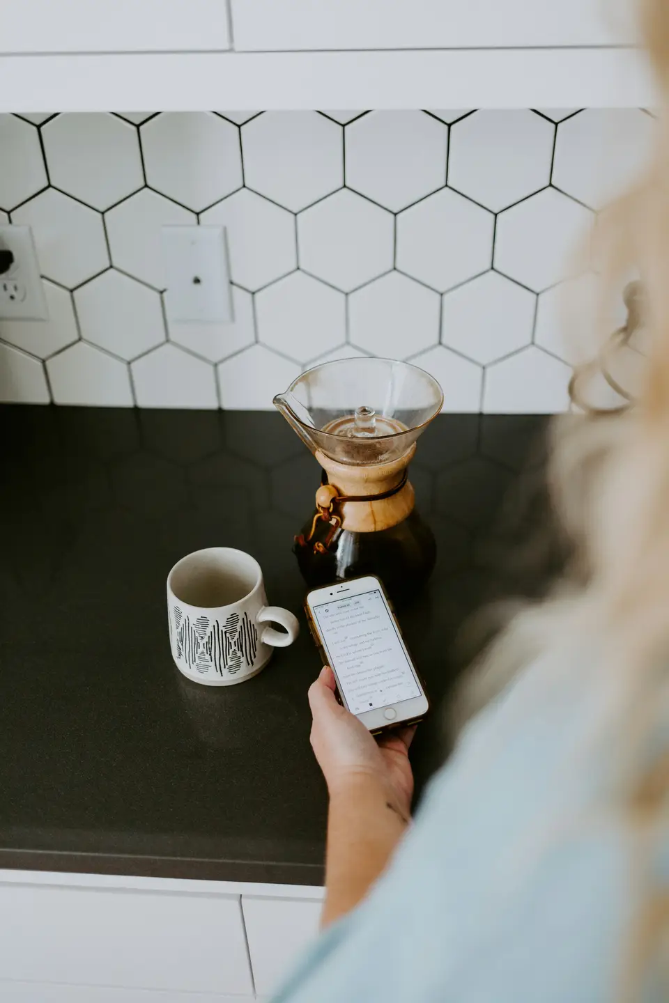 A hand holding a phone above a dark kitchen counter, with a patterned mug and a glass coffee dripper beside it.