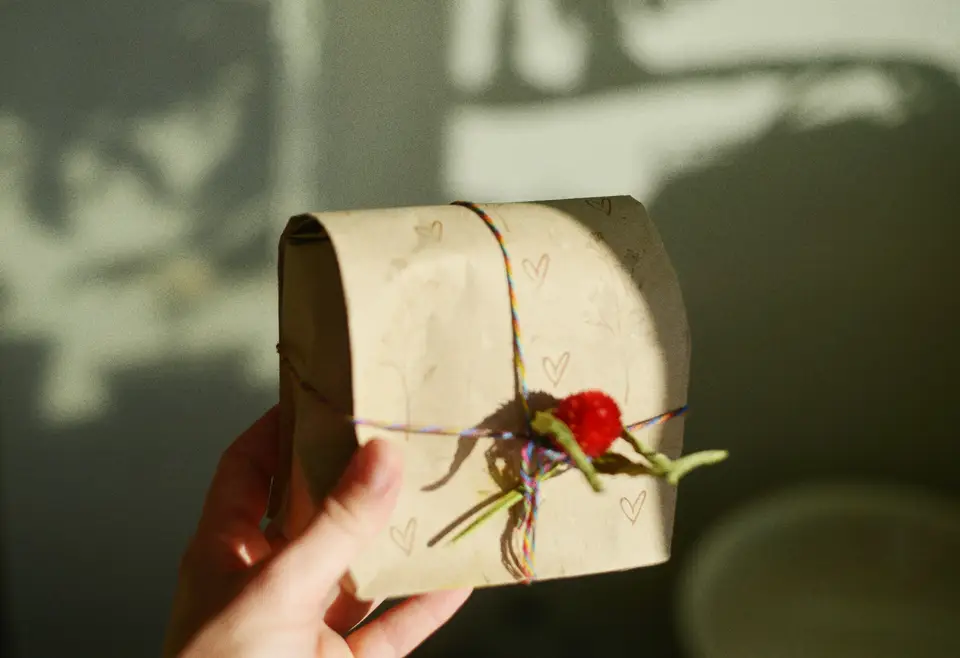 A hand holding a small parcel wrapped in brown paper, tied with colourful string and a red pompom, in late window light.