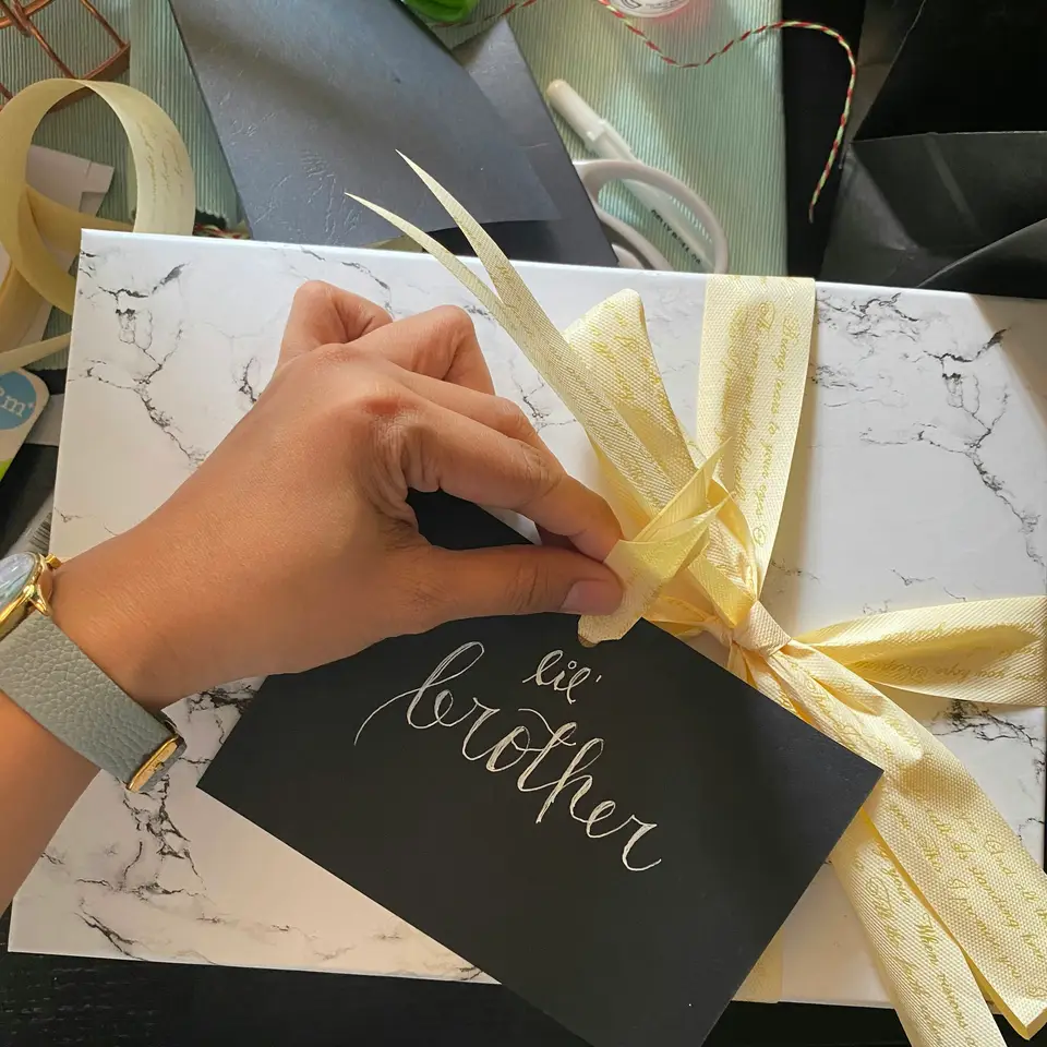 Hands tying a pale yellow ribbon around a gift box on a cluttered table, next to a handwritten card.