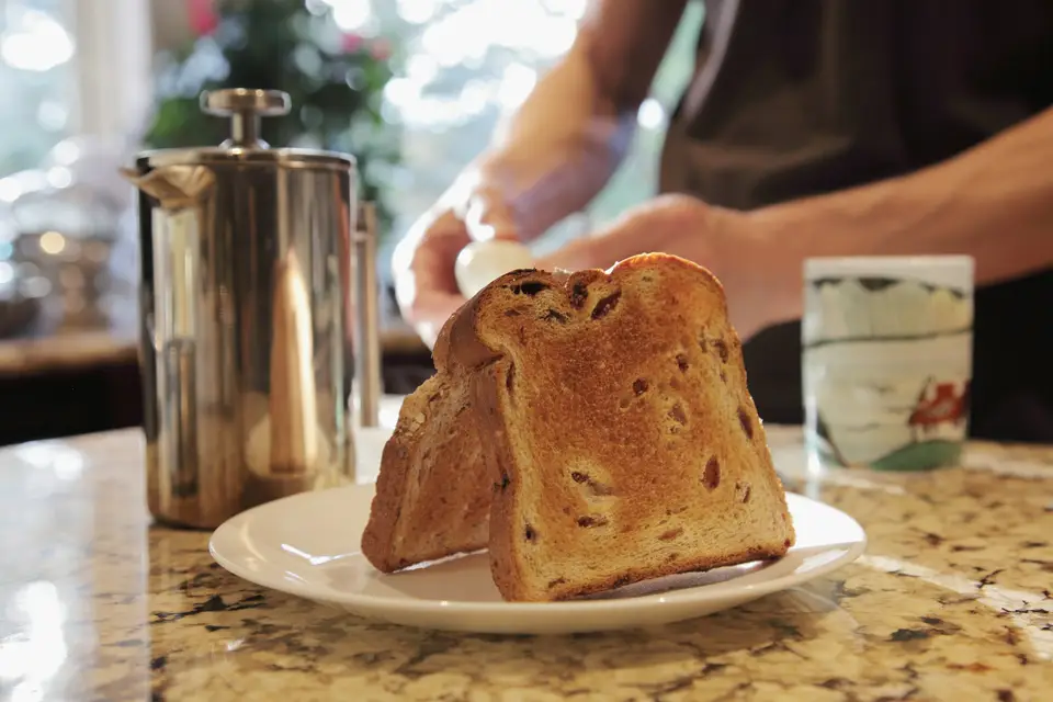 Toast and a glass of milk on a plate at a kitchen counter, with someone's hands visible cracking an egg in the warm morning light behind it.