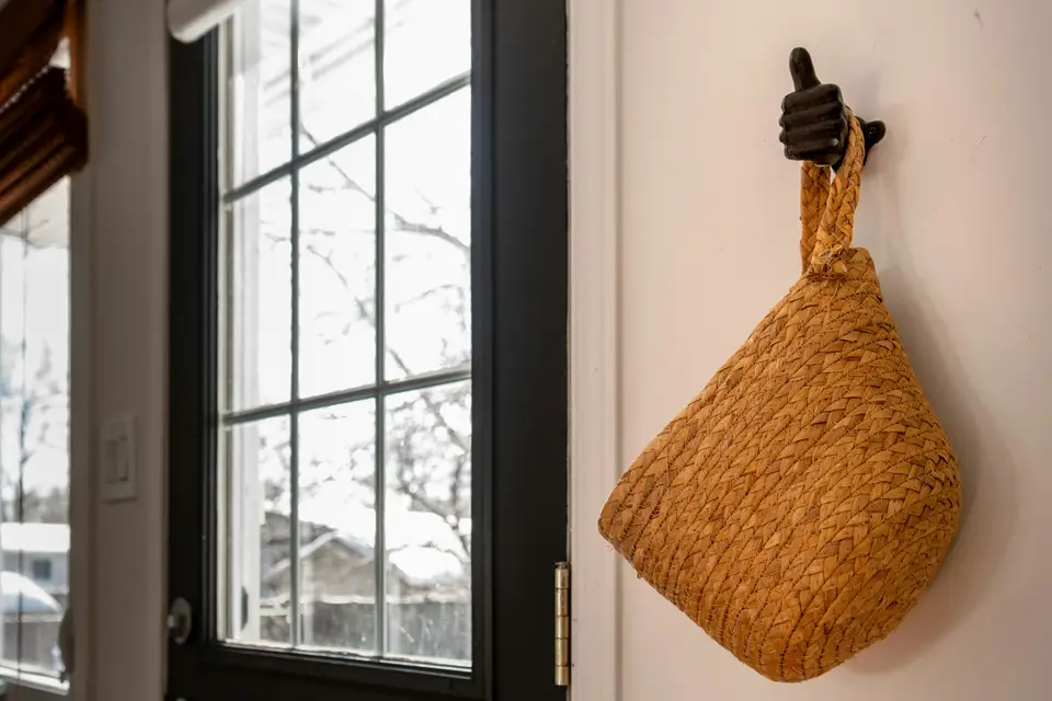 A woven bag hanging from a single hook by a front door, morning light coming through the window beside it.