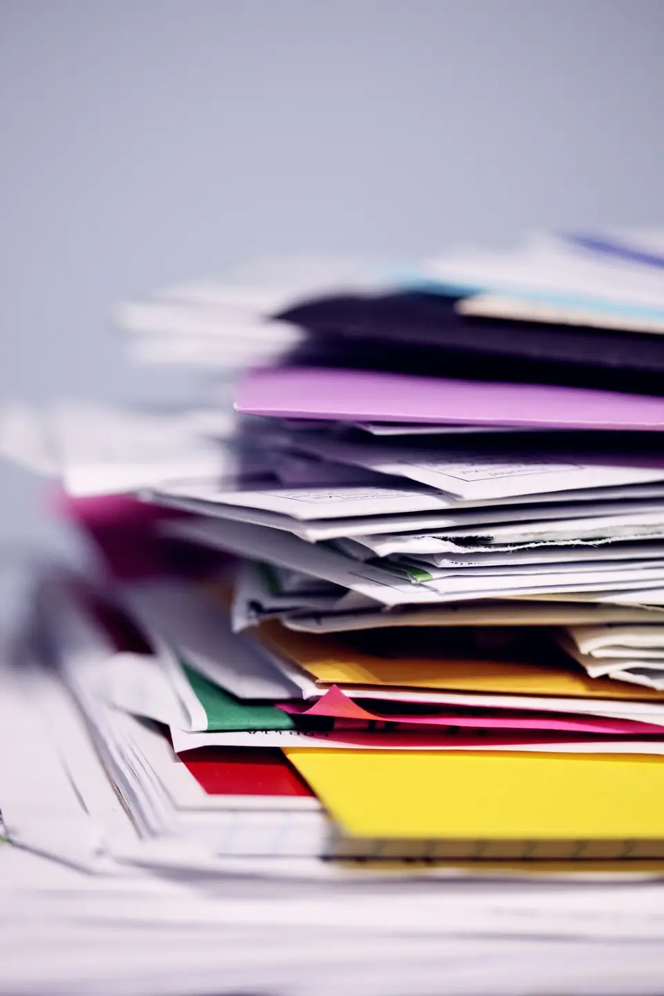 A messy, uneven stack of colorful papers and folders on a blurred surface.