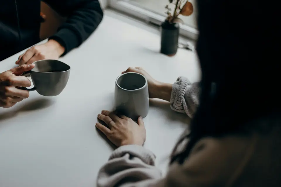 Two people sitting across a pale table, each holding a grey mug, hands resting near a window.