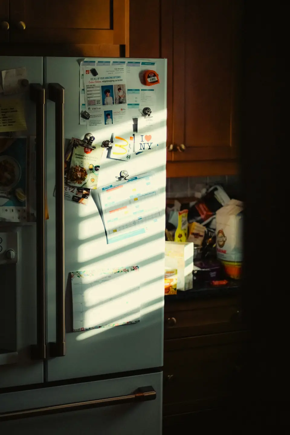 A paper month calendar and school notes held to a fridge door by magnets, striped with light from a window blind.