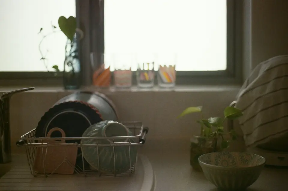 A metal rack of plates and bowls drying beside a sunlit kitchen window, with a striped tea towel hanging at the edge.