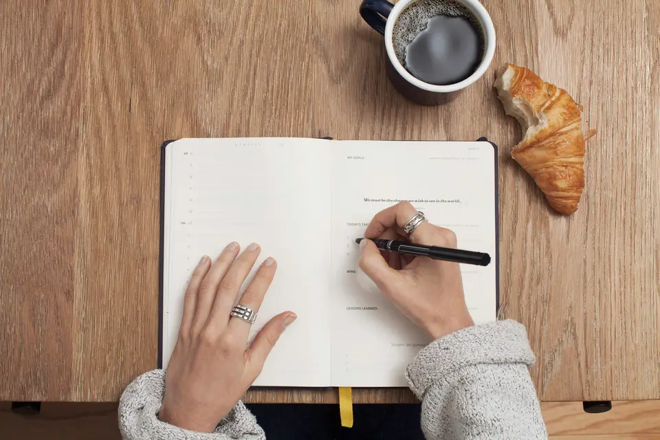 A hand writing in an open notebook on a wooden table, with coffee and a croissant nearby.