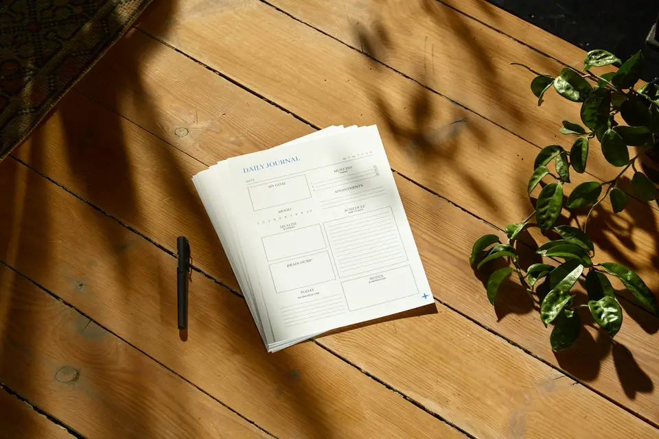 A stack of blank planner sheets and a pen on a sunlit wooden floor, beside a trailing house plant.