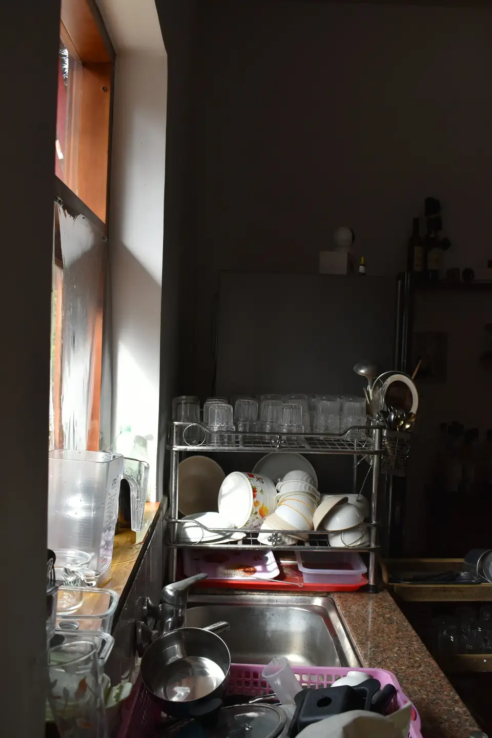 A kitchen in low window light with a drying rack of bowls, a sink full of pans and glasses, and open shelves crowded behind.