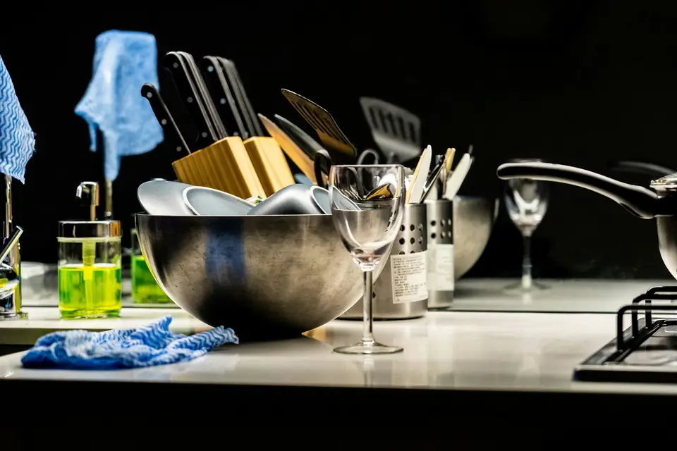A kitchen counter with a steel bowl, a knife block, utensils and a wine glass, a blue cloth lying across the edge.